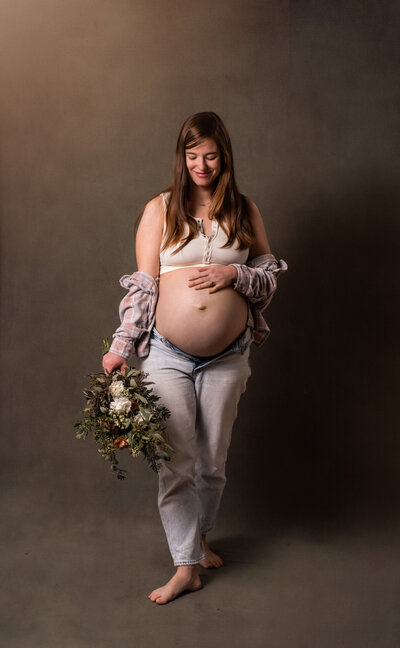 An expecting mama in jeans and a tank top holding flowers and cradling her bump