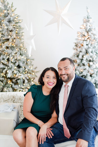 husband and wife sitting on a white couch in front of Christmas Trees by Miami Christmas Mini Session Photographer