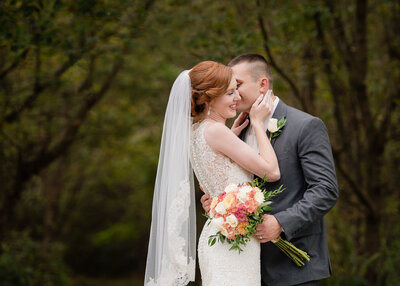 wedding couple bride with red hair smiling while groom kisses her cheek holding floral bouquet