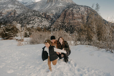 Three people pose in snowy mountains; a child runs toward the camera smiling while a Colorado family photographer capturing real connection kneels behind, documenting this joyful adventure.
