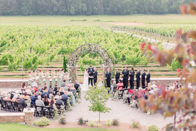 A panoramic view of an outdoor vineyard wedding ceremony with the wedding party and guests gathered around a floral arch, capturing the serene and picturesque landscape.