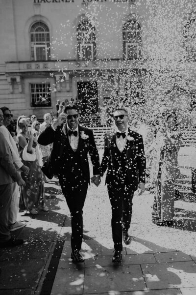 2 grooms walk down the stairs at Hackney Town Hall as their guests throw confetti over them. 