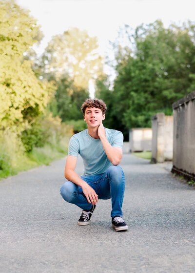 A high school senior in jeans squats in an alley with a hand on his knee