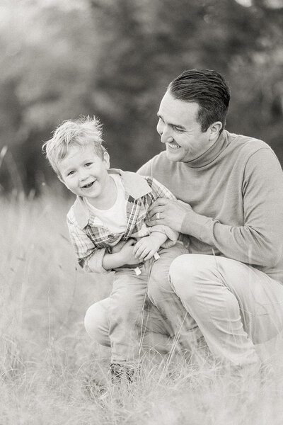 A black and white photo of a dad playing with his young son in a open field at a Dallas park for their family photos.