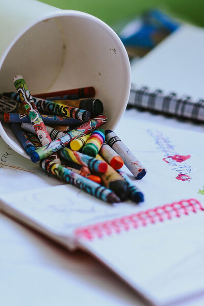 A spilled cup of colorful crayons lies on a table, surrounded by a notebook and sheets of paper with doodles and drawings.