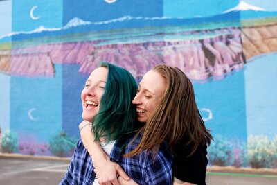 colorful same sex couple laugh and hug in front of bright blue mural