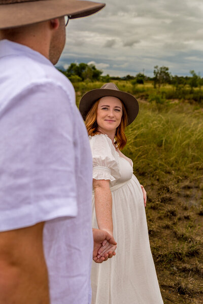 pregnant woman in white dress leading partner through a field - Townsville Maternity Photography by Jamie Simmons