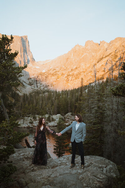 bride and groom are holding hands on a large rock with a lake in the background. trees are surrounding them, and the mountains are turning orange behind them. The bride is in a black dress and the groom is wearing a grey suit. it is sunrise behind the mountains..
