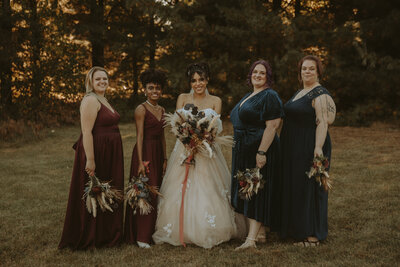 A bride and her bridesmaids giggle in front of the forest while holding their muted, warm-toned bouquets in navy and maroon dresses