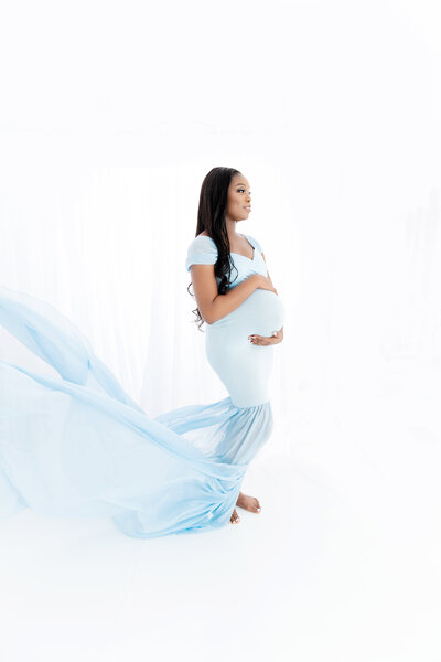 A pregnant woman in a blue maternity gown stands in a studio holding her bump as her train flys behind her thanks to a Greater Atlanta Motherhood & Family Photographer