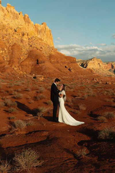 A groom gently holding the face of his bride as they stare into each other's eyes. They are standing in the midst of sagebrush, with red rock and buttes behind them.