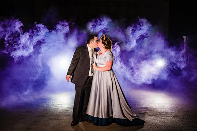 Bride and groom embrace and kiss amidst a purple smoke bomb