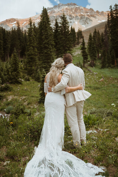 the couple is wearing a wedding dress and a tan suit. they are facing backwards towards a mountain peak. there is greenery surrounding them, and they are embracing each other while enjoying the view. they are eloping in telluride with their dog and finished their hiking elopement in a valley surrounded by the san juan mountain range. they are reading their vows and signing their marriage license in the mountains.