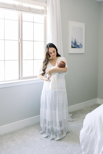 Image of new mother in white dress in home standing next to window holding baby taken by maternity photographer sacramento Kelsey Krall