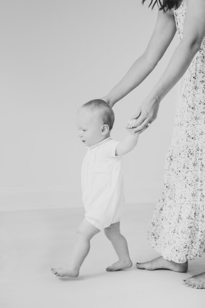 Black and white image of baby boy walking in white romper as mom in blue dress holds his hands