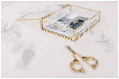 Family session photos in a glass and gold box with a pair of gold scissors and blue ribbon nearby