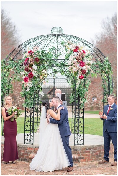 Bride and groom standing in front of a pavilion with florals having their first kiss.