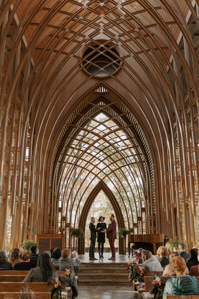 A gay couple stands at the altar of Cooper Memorial Chapel in Bella Vista, Arkansas as they get married on Halloween. Their officiant is dressed in a solid black dress with a witch hat. Their friends and family watch as the couple marries from the pews.
