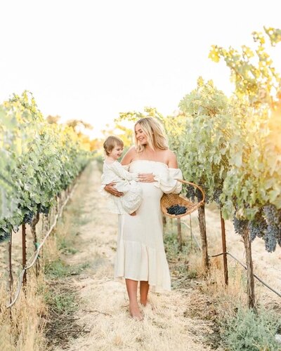 A maternity photography session by Bay area photographer shows a mother holding  and walking with her toddler daughter while caressing her baby bump in an orchard.