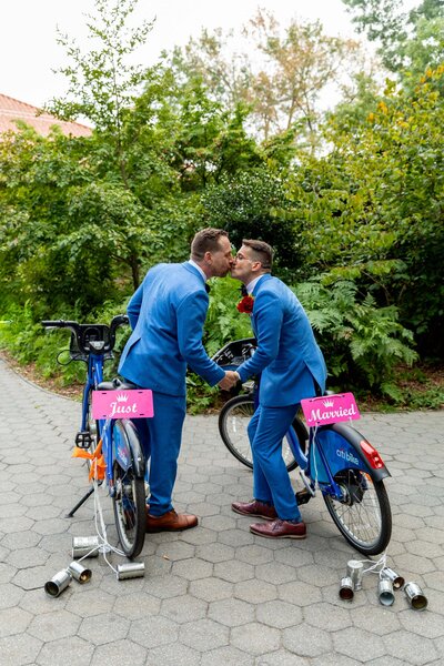 Two grooms on bikes leaning towards each other to kiss.