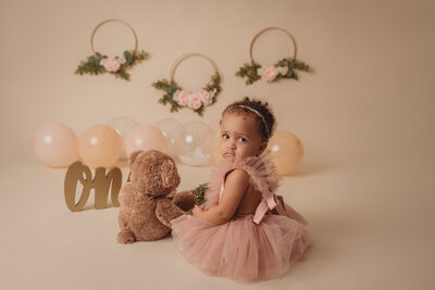 Beautiful modern cake smash portrait with one year old girl wearing pink tulle dress at atlanta milestone photography studio sitting next to teddy bear with white cake, roses and balloons in background