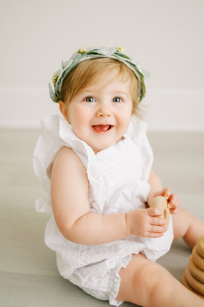 One year old baby girl wearing white romper and floral headband smiles during baby photoshoot