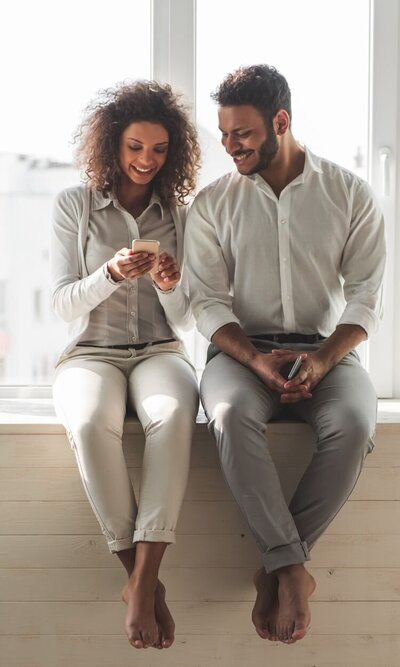 A couple sitting on a bench as they smile at something on the woman's phone. This could symbolize a couple searching options for online couples therapy in Florida. Contact a marriage counselor for support with online marriage counseling and other services.
