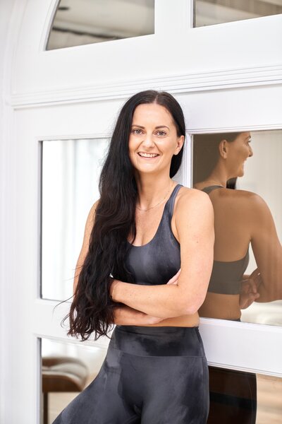 Yoga instructor Sarah smiles confidently, standing by a window in a bright studio, reflecting an inviting and positive atmosphere for wellness and personal growth.