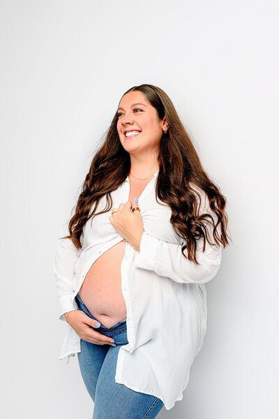 Pregnant mother wearing jeans and a white button down shirt with her expectant belly showing standing against a studio white wall smiling off camera
