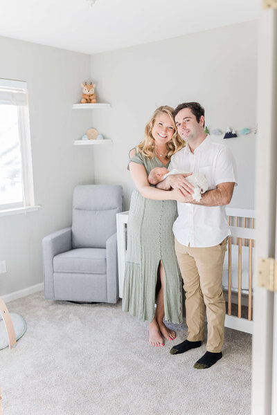 Mom and Dad standing together in boho styled nursery. They are standing in front of a white crib and Dad is holding newborn baby boy whose swaddled and sleeping