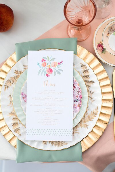 table setting at a wedding at Ravenswood Mansion with a fluted gold charger, a vintage china dinner plate, vintage china salad plate and a vintage china dessert plate accented by a teal napkin and peach floral menu card on a peach and white striped table cloth