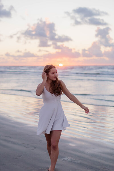 Highschool senior graduating soon walks towards camera on Galveston Beach at sunrise in white dress