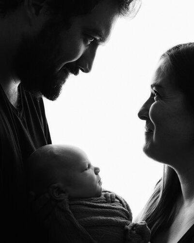 Black silhouette on white background of a mother and father facing each other holding their newborn and smiling