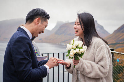 A Chinese couple is exchanging rings during their elopement ceremony in Senja, Norway, with a dramatic mountain range and fjord in the background. The man, wearing a navy blue suit is visibly excited, as he slides the ring onto the woman's finger. The woman, in a beige, fluffy coat and smiling joyfully, is holding a bouquet of white roses. The overcast and rainy sky and autumn colours in the background adds to the dramatic atmosphere of the occasion.