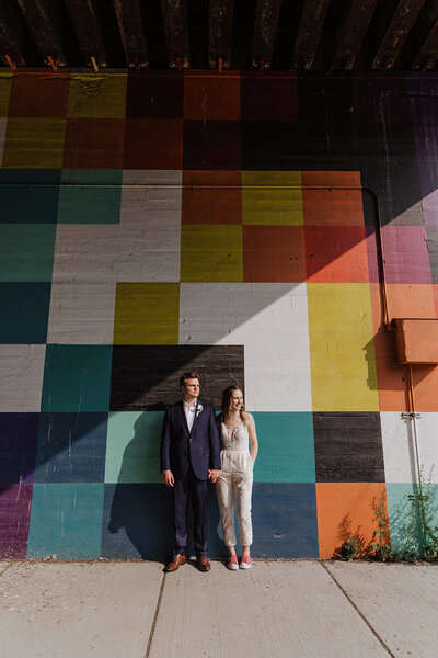 wedding couple leans against colorful mural in Fulton Market Chicago. She wears a bridal jumpsuit and he is in a navy suit.