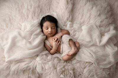 sleeping newborn baby boy with a lot of dark hair wearing white knit overalls laying on cream flokati rug