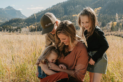A woman and three children smile together in a sunny field with mountains in the background, capturing the warmth of candid family photography at golden hour.