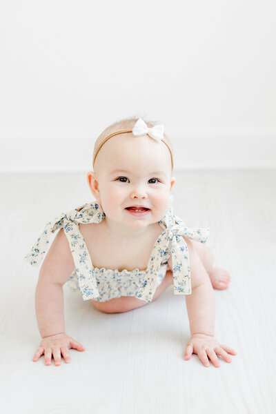 6 month old baby girl smiles while crawling in a floral romper and white bow headband