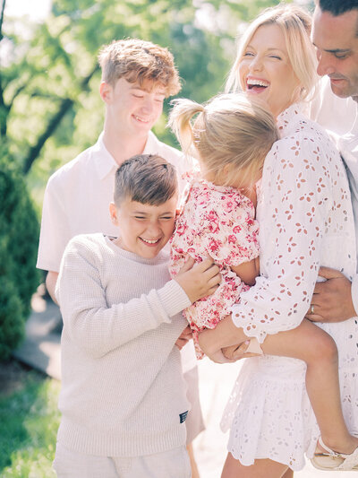 Blonde mother holds her little girl as her two sons and husband tickle them during their family photo session.