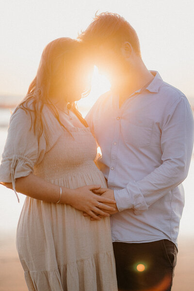 A maternity photoshoot photo with the sunset behind the couple with their hands on the baby bump, photographed by Tashina Narelle Photography