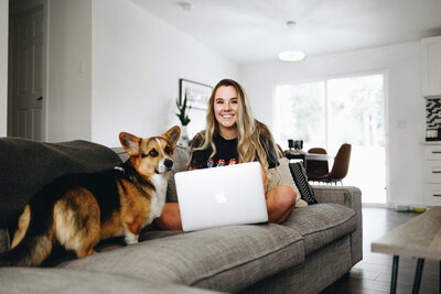 Showit websites designer, Clair Schwem, working on her couch on a luxury branding project. She is sitting on a grey couch. A tri-color corgi, Cannon, is sitting to her left.