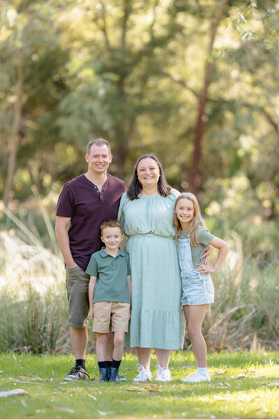 Outdoor family portrait in Geelong with parents and their two children smiling together in a sun-dappled bushland reserve.
