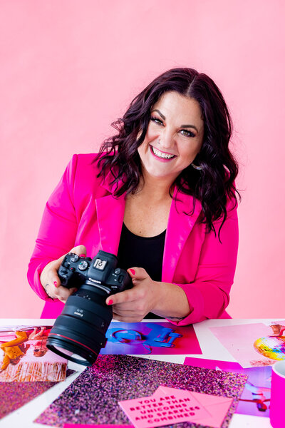 Brand photo of woman in hot pink jacket holding a Canon Camera with glitter paper on her desk and a light pink backdrop behind her