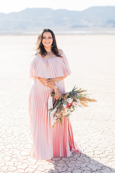 New Jersey photographer, Kiamarie Stone, smiles for photo in the desert holding a bouquet in pink full length gown