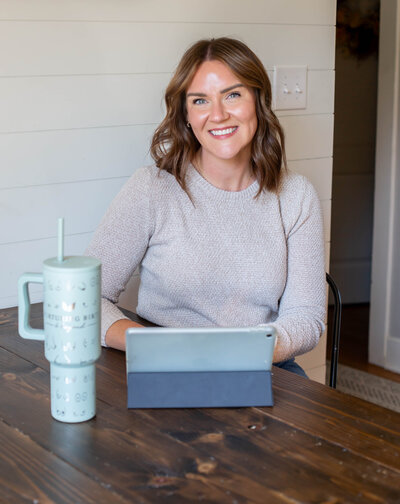 In-Home lactation consultant, Sarah, sitting a a wooden desk with an iPad and tumbler wearing a cream colored sweater