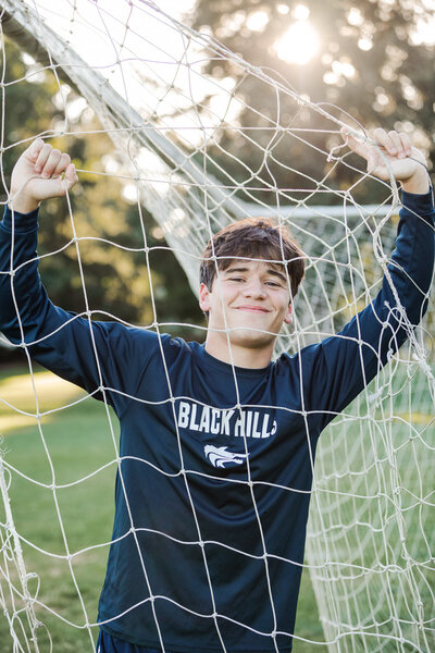 Older teenager male, posing with arms up holding onto a soccer net. smiling with the sun shining behind him