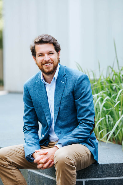 Portrait of a brown-haired bearded man sitting on a marble bench. He's wearing a blue wool blazer, a white dress shirt and khaki pants.