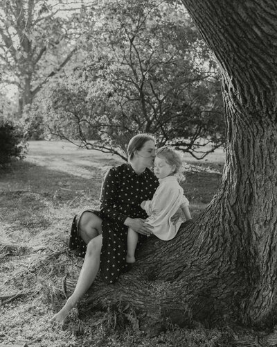 Perhekuvaus äidistä ja hänen 3-vuotiaasta pojastaan istumassa puunrungolla äidin antaessa suukko pojan ohimolle puistossa Helsingissä