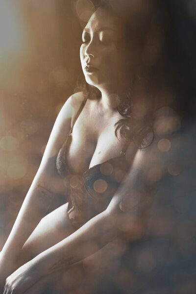 An artistic boudoir photo captures a woman from the side and partially from above as she sits in a San Fransisco studio.She's adorned in a matching set of bra and underwear with light illuminating her from behind. The image is enhanced with an artistic overlay, adding a creative and ethereal quality to the portrait.
