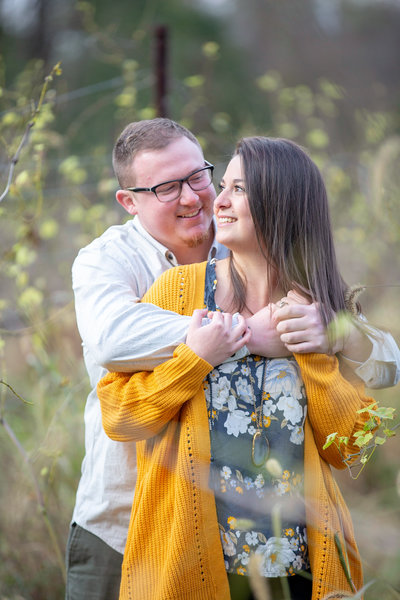 fiance embraces his soon-to-be bride from behind in a field during their engagement photography session